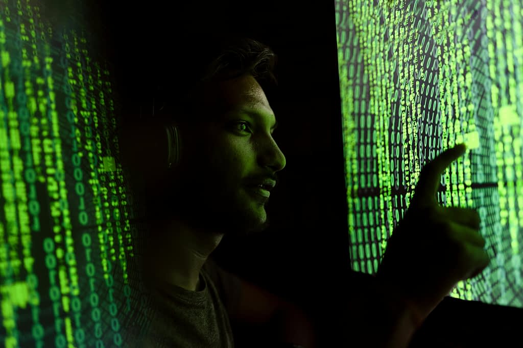 A man is standing in the dark in between two huge shining screens showing lines of code in green on it. The man is touching one of the screens.